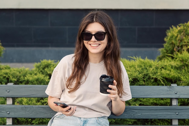 Eine junge Frau sitzt mit einer Tasse Kaffee und einem Smartphone auf einer Bank