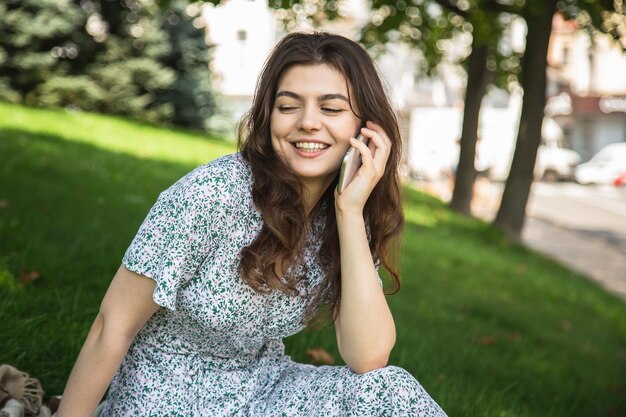 Eine junge Frau spricht auf einem Smartphone, während sie auf dem Gras im Park sitzt