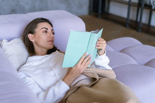 Eine junge Frau zu Hause in einem weißen Hoodie auf der Couch hüllt sich in eine warme Decke, liest ein Buch