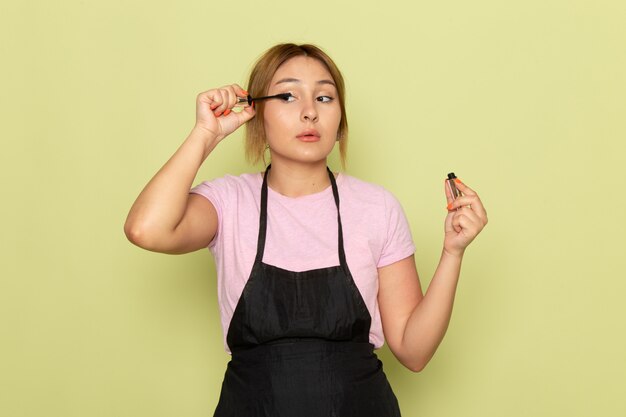 Eine junge Friseurin der Vorderansicht im rosa T-Shirt und im schwarzen Umhang, der Make-up auf Grün tut