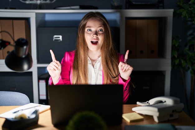 Eine junge kaukasische Frau, die nachts im Büro arbeitet, blickt erstaunt und überrascht auf und zeigt mit Fingern und erhobenen Armen