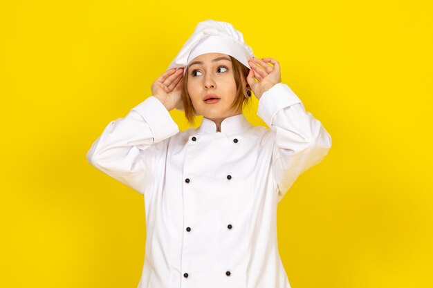 Eine junge Köchin der Vorderansicht im weißen Kochanzug und in der weißen Kappe, die ihren Anzug auf dem Gelb fixiert