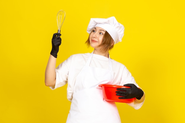 Eine junge Köchin der Vorderansicht im weißen Kochanzug und in der weißen Kappe in den schwarzen Handschuhen, die rote Schüssel halten, die es auf dem gelben mischt