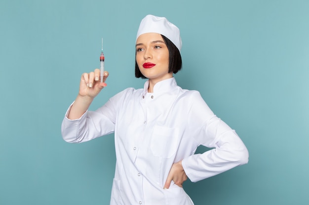Eine junge Krankenschwester der Vorderansicht im weißen medizinischen Anzug, der die Injektion auf dem blauen Schreibtischmedizin-Krankenhausarzt vorbereitet