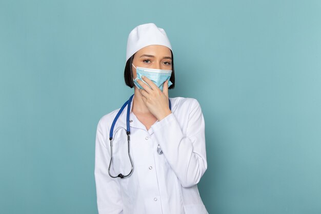 Eine junge Krankenschwester der Vorderansicht im weißen medizinischen Anzug und im blauen Stethoskop, das Maske auf dem blauen Schreibtischmedizin-Krankenhausarzt trägt