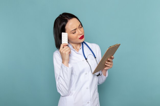 Eine junge Krankenschwester der Vorderansicht im weißen medizinischen Anzug und im blauen Stethoskop, das Notizblock hält