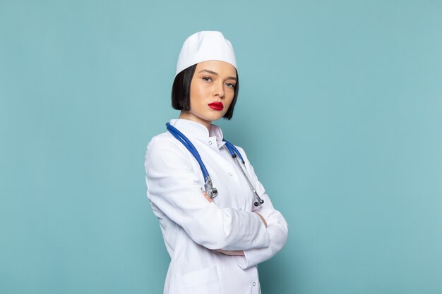 Eine junge Krankenschwester der Vorderansicht im weißen medizinischen Anzug und im blauen Stethoskop, die auf dem medizinischen Krankenhausarzt des blauen Schreibtisches aufwerfen