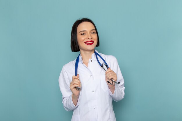 Eine junge Krankenschwester der Vorderansicht im weißen medizinischen Anzug und im blauen Stethoskop lächelnd auf dem blauen Schreibtischmedizinkrankenhausarzt
