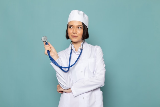 Eine junge Krankenschwester der Vorderansicht im weißen medizinischen Anzug und im blauen Stethoskop mit denkendem Ausdruck auf dem blauen Schreibtischmedizin-Krankenhausarzt