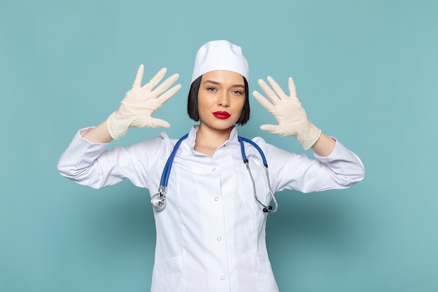 Eine junge Krankenschwester der Vorderansicht im weißen medizinischen Anzug und im blauen Stethoskop zeigen ihre Hände auf dem blauen Schreibtischmedizin-Krankenhausarzt