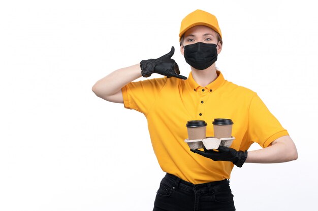 Eine junge Kurierin der Vorderansicht in den gelben einheitlichen schwarzen Handschuhen und in der schwarzen Maske, die Kaffeetassen halten, die Befehl liefern, um zu bitten, anzurufen