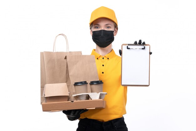 Eine junge Kurierin der Vorderansicht in den gelben einheitlichen schwarzen Handschuhen und in der schwarzen Maske, die Pizzaschachteln und den Notizblock halten liefert
