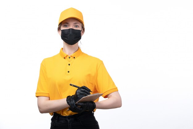 Eine junge Kurierin der Vorderansicht in den schwarzen Handschuhen der gelben Uniform und der schwarzen Maske, die den Notizblock hält, der Aufträge aufschreibt