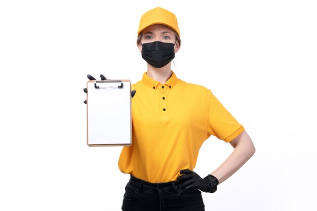 Eine junge Kurierin der Vorderansicht in den schwarzen Handschuhen der gelben Uniform und der schwarzen Maske, die Notizblock hält und zeigt