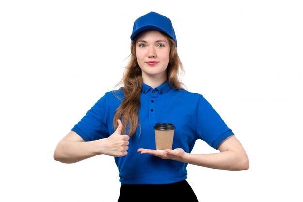Eine junge Kurierin der Vorderansicht in der blauen Kappe des blauen Hemdes und der schwarzen Hose lächelnd, die Kaffeetasse auf Weiß hält