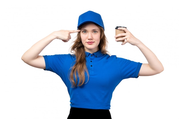 Eine junge Kurierin der Vorderansicht in der blauen Kappe des blauen Hemdes und in der schwarzen Hose, die Kaffeetasse hält, die auf ihren Kopf auf Weiß zeigt