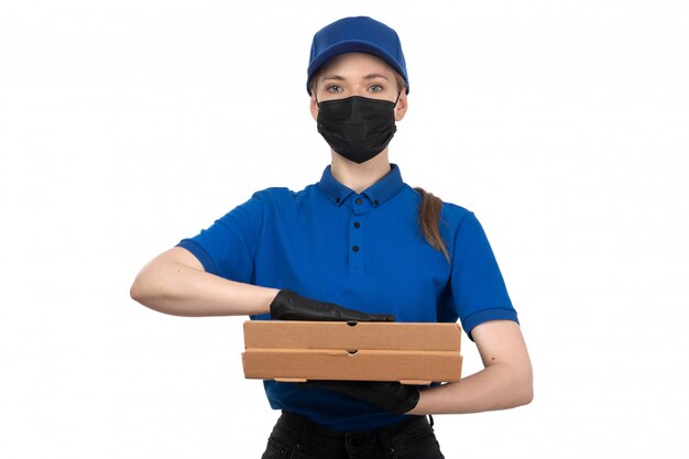 Eine junge Kurierin der Vorderansicht in der blauen Maske der blauen Uniform und in den Handschuhen, die Lebensmittel-Lieferpakete halten