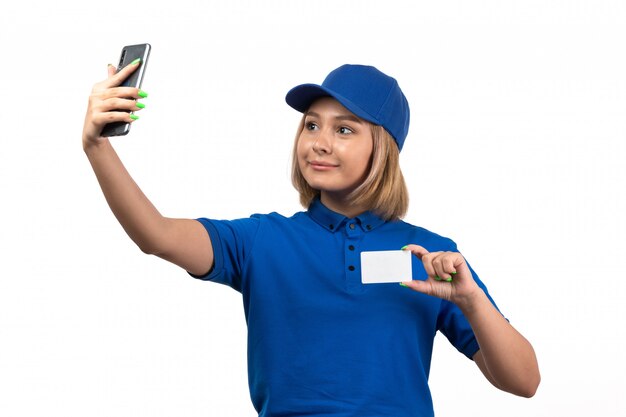 Eine junge Kurierin der Vorderansicht in der blauen Uniform, die Telefon und weiße Karte hält, die ein selfie nehmen