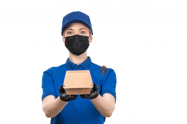 Eine junge Kurierin der Vorderansicht in der blauen Uniformschwarzmaske und in den Handschuhen, die Lebensmittel-Lieferpaket halten