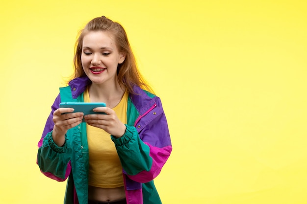 Eine junge moderne Frau der Vorderansicht in der schwarzen Hose des gelben Hemdes und der bunten Jacke, die etwas am Telefon glücklichen Ausdruck beobachten