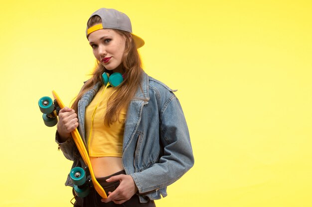 Eine junge moderne Frau der Vorderansicht in der schwarzen Hose des gelben Hemdes und im Jeansmantel, der Skateboard mit farbigen Kopfhörern hält