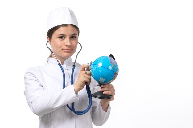 Eine junge Ärztin der Vorderansicht im weißen medizinischen Anzug mit blauem Stethoskop, das Globus prüft