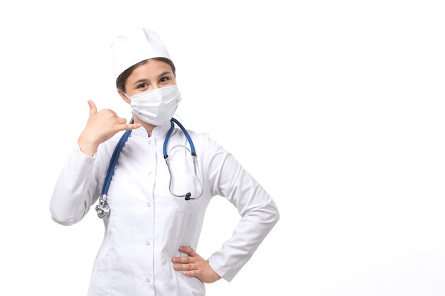 Eine junge Ärztin der Vorderansicht im weißen medizinischen Anzug mit blauem Stethoskop, das weiße Maske trägt