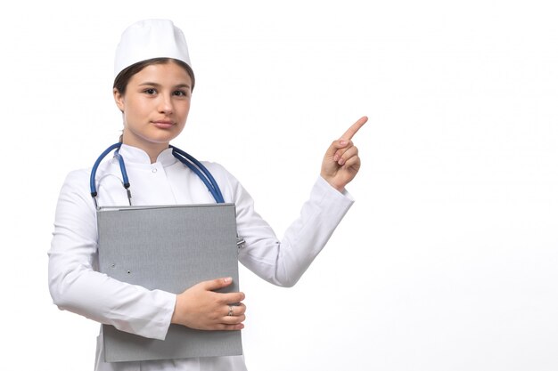 Eine junge Ärztin der Vorderansicht im weißen medizinischen Anzug mit blauen Stethoskop, das Dokumente hält