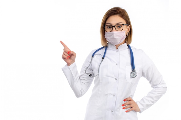 Eine junge Ärztin der Vorderansicht im weißen medizinischen Anzug mit Stethoskop, das weiße Schutzmaske auf dem Weiß trägt