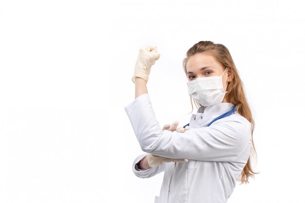 Eine junge Ärztin der Vorderansicht im weißen medizinischen Anzug mit Stethoskop, das weiße Schutzmaske in Handschuhen trägt, die auf dem Weiß biegen
