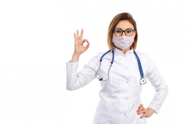 Eine junge Ärztin der Vorderansicht im weißen medizinischen Anzug mit Stethoskop, das weiße Schutzmaske trägt, die darstellendes gutes Zeichen auf dem Weiß aufwirft