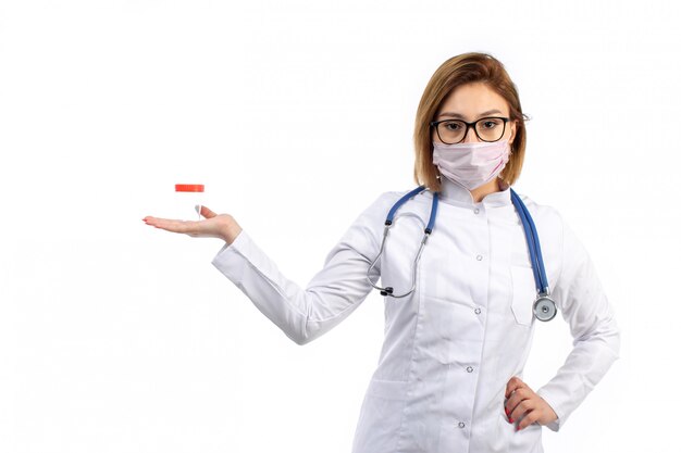 Eine junge Ärztin der Vorderansicht im weißen medizinischen Anzug mit Stethoskop, das weiße Schutzmaske trägt, die Flasche auf dem Weiß hält