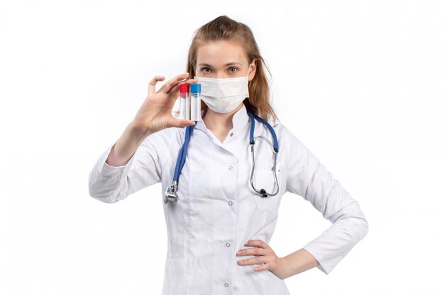 Eine junge Ärztin der Vorderansicht im weißen medizinischen Anzug mit Stethoskop, das weiße Schutzmaske trägt, die Halteflaschen auf dem Weiß aufwirft