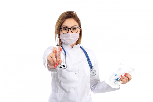 Eine junge Ärztin der Vorderansicht im weißen medizinischen Anzug mit Stethoskop, das weiße Schutzmaske trägt, die Injektion auf dem Weiß hält