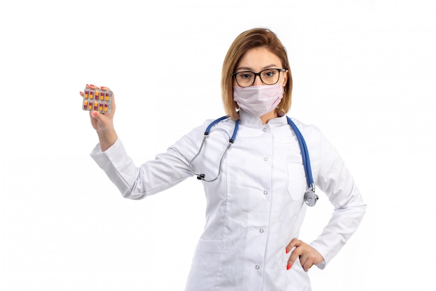 Eine junge Ärztin der Vorderansicht im weißen medizinischen Anzug mit Stethoskop, das weiße Schutzmaske trägt, die Pillen auf dem Weiß hält