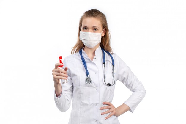 Eine junge Ärztin der Vorderansicht im weißen medizinischen Anzug mit Stethoskop, das weiße Schutzmaske trägt, die Spray auf Weiß hält