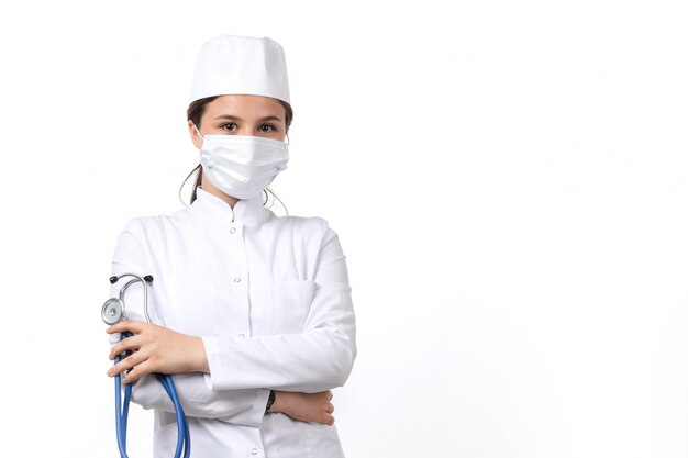 Eine junge Ärztin der Vorderansicht im weißen medizinischen Anzug und in der weißen Kappe mit blauem Stethoskop, das eine Maske trägt