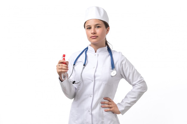 Eine junge Ärztin der Vorderansicht im weißen medizinischen Anzug und in der weißen Kappe mit blauem Stethoskop, das Spray hält