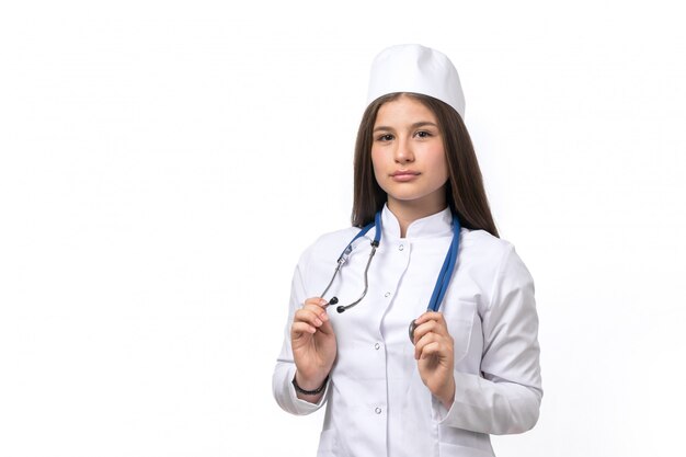 Eine junge Ärztin der Vorderansicht im weißen medizinischen Anzug und in der weißen Kappe mit dem blauen Stethoskop, das aufwirft