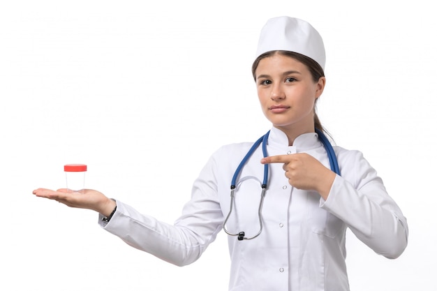 Eine junge Ärztin der Vorderansicht im weißen medizinischen Anzug und in der weißen Kappe mit dem blauen Stethoskop, das Flasche hält