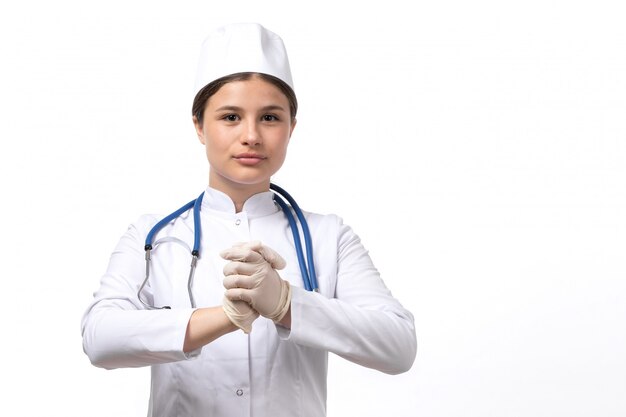 Eine junge Ärztin der Vorderansicht im weißen medizinischen Anzug und in der weißen Kappe mit dem blauen Stethoskop, das Handschuhe trägt