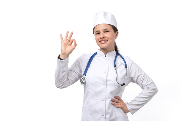 Eine junge Ärztin der Vorderansicht im weißen medizinischen Anzug und in der weißen Kappe mit dem lächelnden blauen Stethoskop