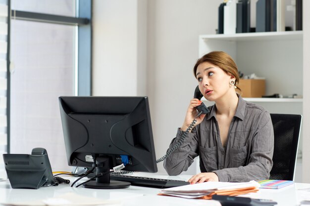 Eine junge schöne Dame der Vorderansicht im grauen Hemd, das an ihrem PC arbeitet, der in ihrem Büro sitzt, das am Stadttelefon während der Tagesaufbaujobaktivität spricht