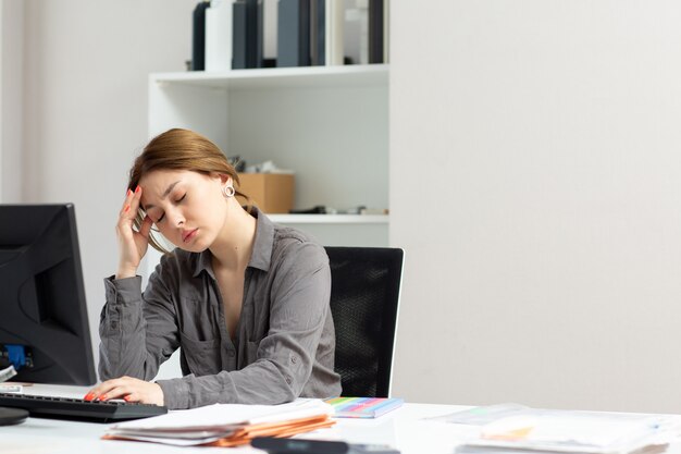 Eine junge schöne Dame der Vorderansicht im grauen Hemd, das mit den Dokumenten arbeitet, die ihren PC verwenden, der in ihrem Büro sitzt, das unter Kopfschmerzen während der Tagesaufbau-Jobaktivität leidet