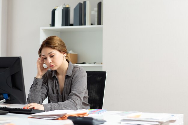 Eine junge schöne Dame der Vorderansicht im grauen Hemd, das mit den Dokumenten arbeitet, die ihren PC verwenden, der in ihrem Büro sitzt und denkt, während des Tagesaufbaus Jobaktivität besorgniserregend