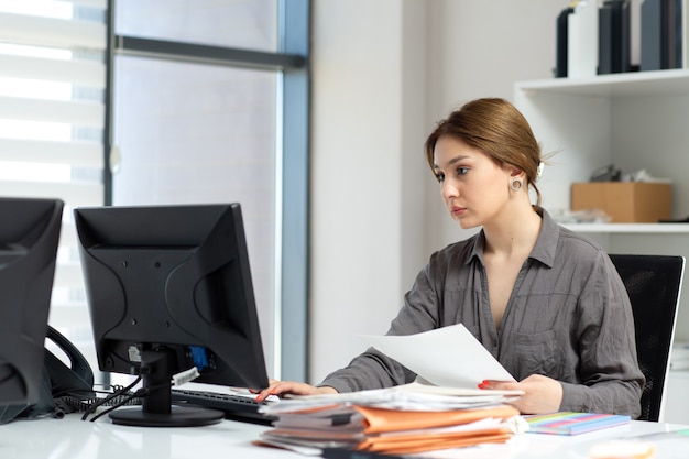 Eine junge schöne Dame der Vorderansicht im grauen Hemd, das mit den Dokumenten und dem Laptop arbeitet, die in ihrem Büro während der Tagesaufbauarbeitstätigkeit sitzen