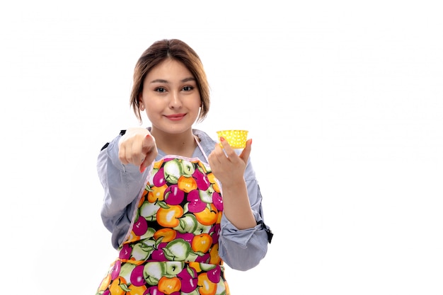 Eine junge schöne Dame der Vorderansicht im hellblauen Hemd und im bunten Umhang, der gelben Kuchenpfannen glücklich lächelnden Ausdruck hält