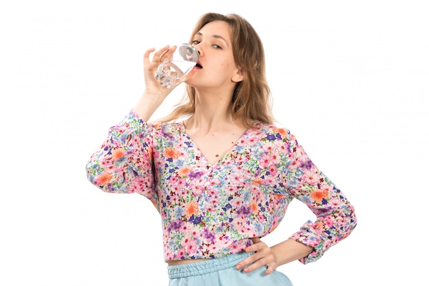 Eine junge schöne Dame der Vorderansicht im Hemd der bunten Blume und im blauen Rock, der Wasser auf dem Weiß trinkt