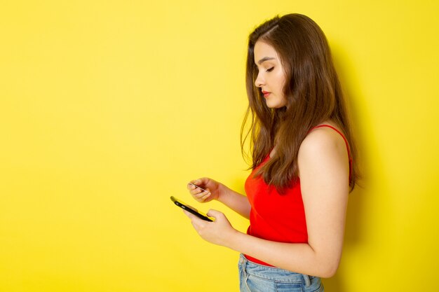 Eine junge schöne Dame der Vorderansicht im roten Hemd und in den blauen Jeans unter Verwendung eines Telefons