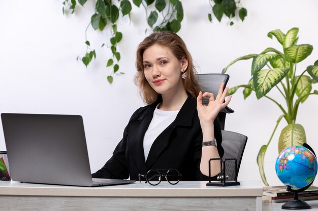 Eine junge schöne Dame der Vorderansicht im weißen Hemd und in der schwarzen Jacke, die ihren Laptop vor dem lächelnden Tisch zeigt, der Zeichen mit hängenden Blättern zeigt